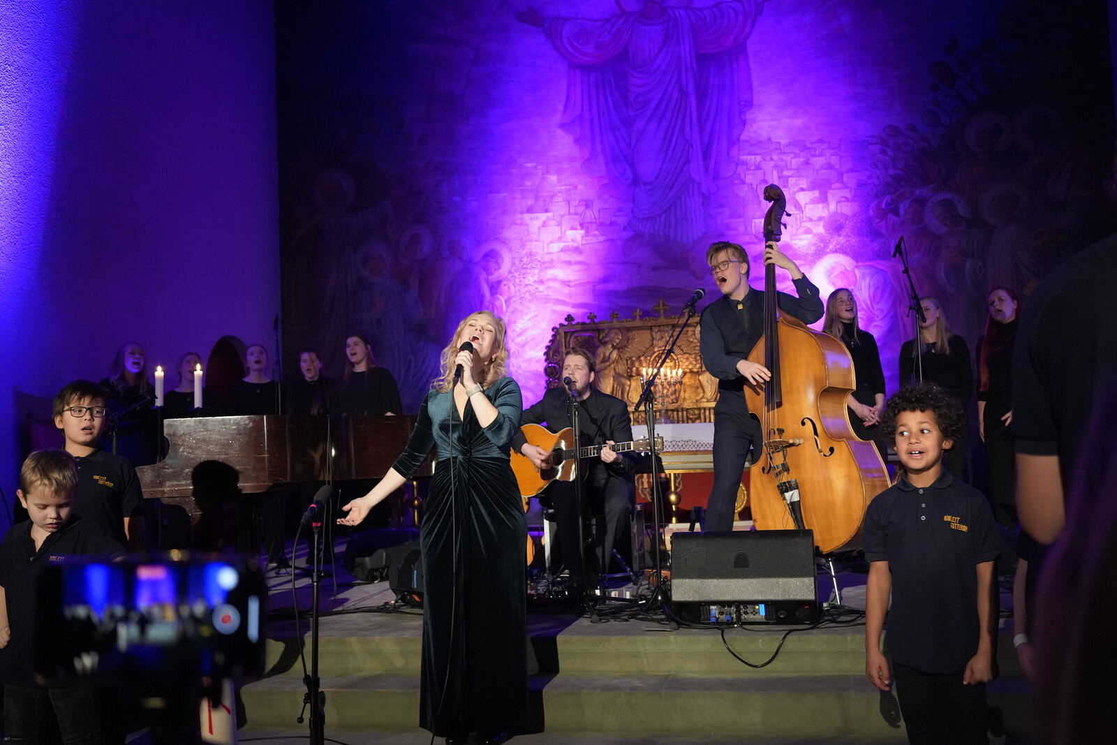 Kristin Minde synger i en kirke med barnekor, gitarist og kontrabassist