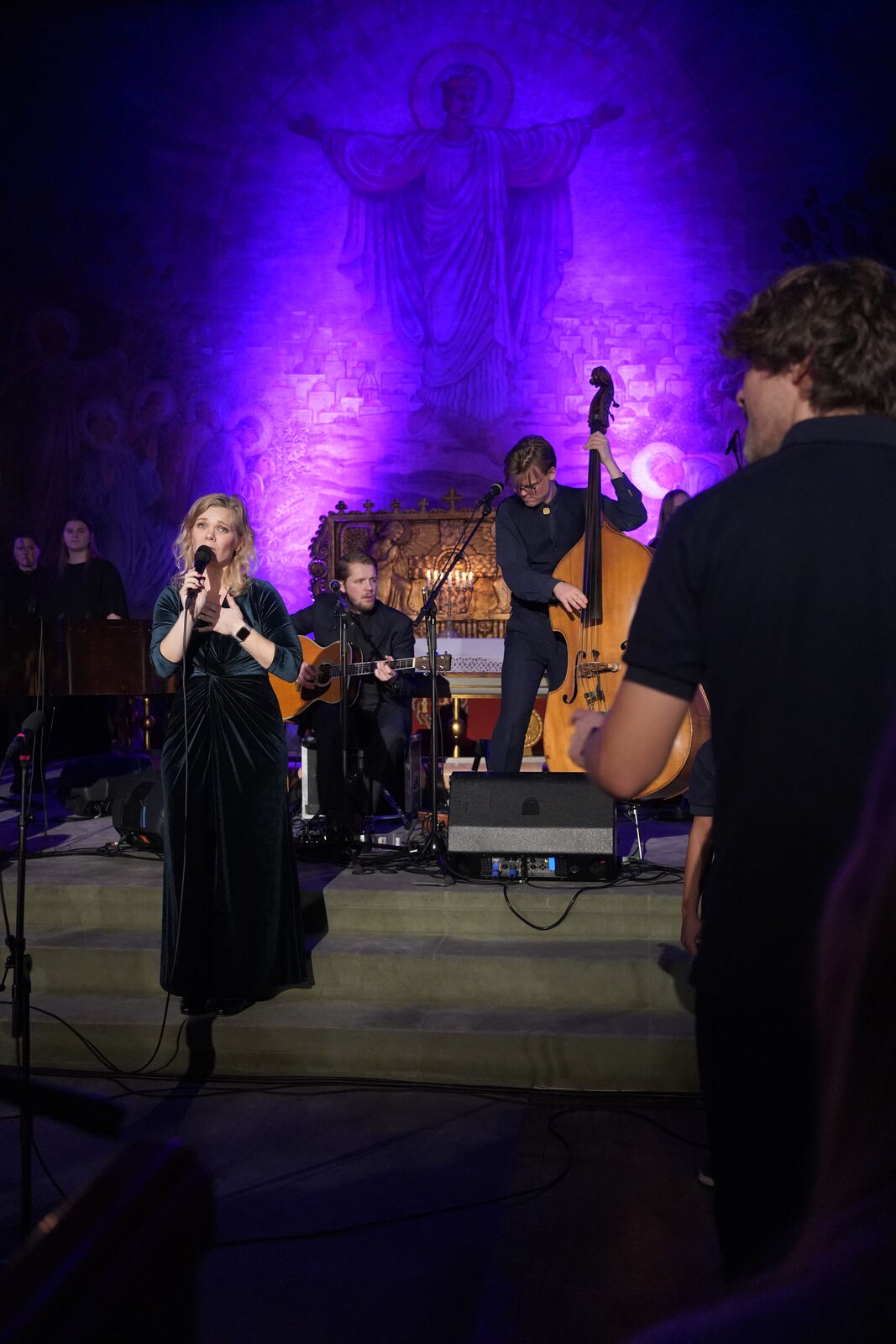 Kristin Minde synger på scenen i en kirke opplyst i lilla, med band og kor bak seg